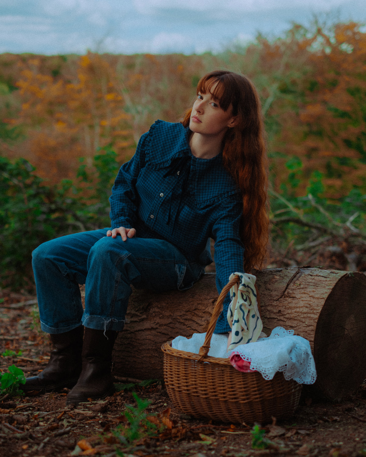 A woman with long red hair sits on a log outdoors, wearing a dark blue patterned button-up shirt and blue jeans. A wicker picnic basket with a white towel and a patterned mug rests beside her on the forest floor, with autumn foliage in the background.